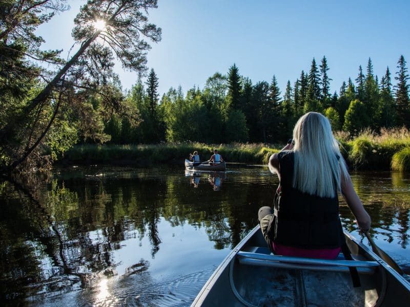 Blond kvinna paddlar kanot på ett spegelblankt vatten under en blå himmel