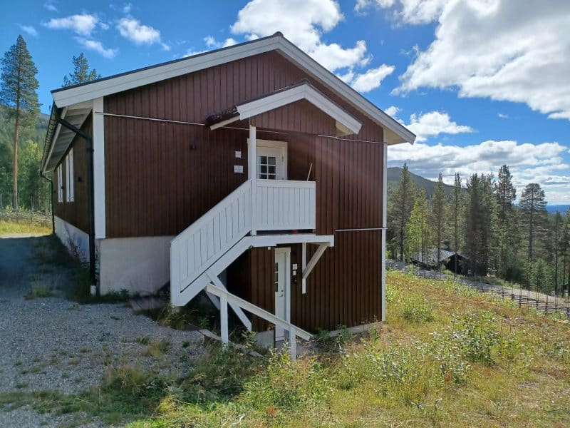 Björnbyn, brown wooden building with white stair railing on a verdant mountain slope with a blue summer sky in the background.