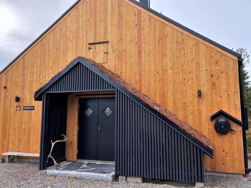 Modern mountain house. Asymmetric light wood gable with a black painted asymmetric porch with sedum roof housing the front door and a ski storage room. Against the wall is a rustic wooden bench. On the wall is the number 23. There is also a hatch on the wall above the veranda. At the side of the veranda is a charger for electric cars. In front of the doors is a stone paving in slate. A reindeer antler leans against the wall in the veranda.