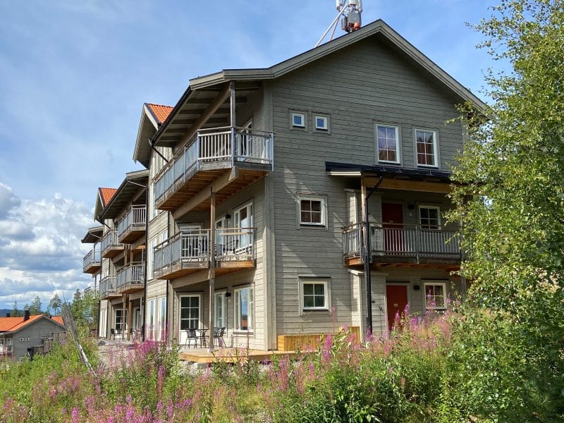 Exteriörbild av grå träfastighet med lägenheter på skalshöjden. Huset står på en sluttning med blå himmel i bakgrunden, omgiven av lummiga ljusgröna björkar.