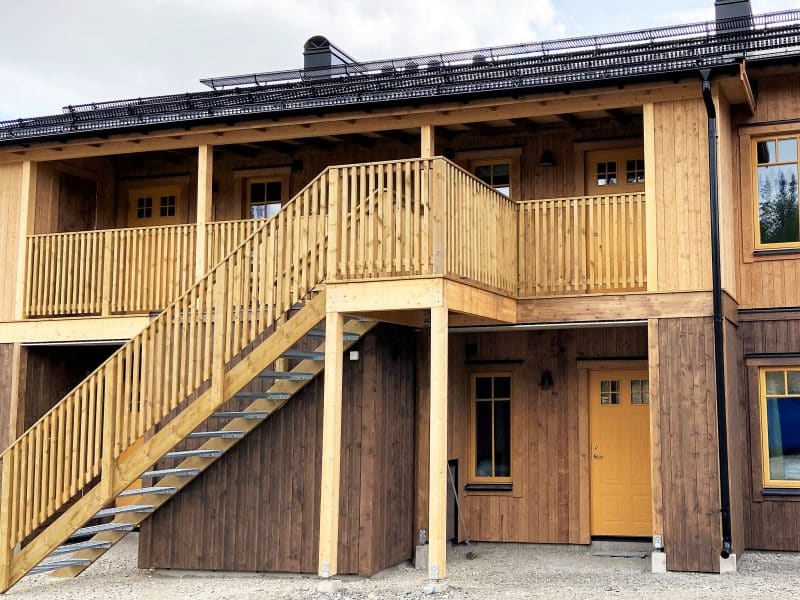 Exterior of apartment living in Vemdalsporten. Wooden property on two floors with a facade that is stained in a darker color on the ground floor and wood-colored on the upper floor, as well as the wooden railing on the stairs up to the upper floor. Front door, window frames and rafters are yellow.